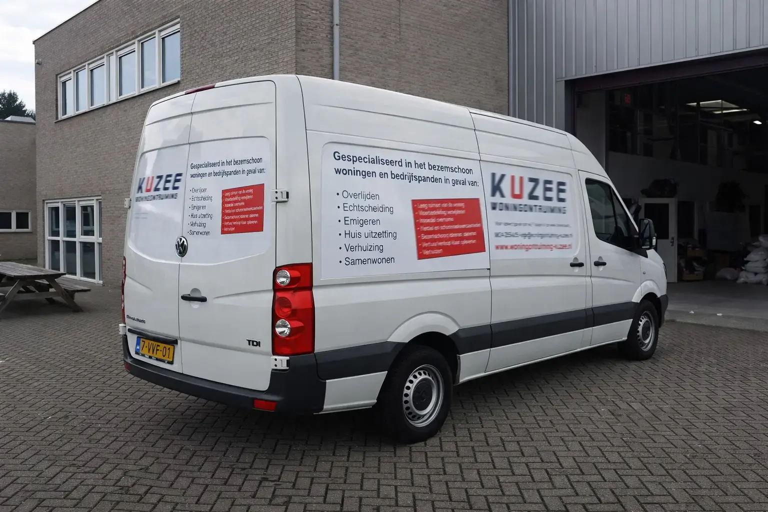 Witte bestelbus met full-colour voertuigbelettering van KUZEE Woningontruiming, voorzien van logo, dienstenoverzicht en rode tekstvlakken, geparkeerd bij een bedrijfspand.